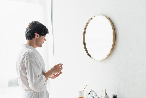 young man in white bathrobe holding cosmetic cream near mirror in bathroom