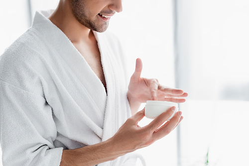 cropped view of man in white bathrobe holding container with cosmetic cream