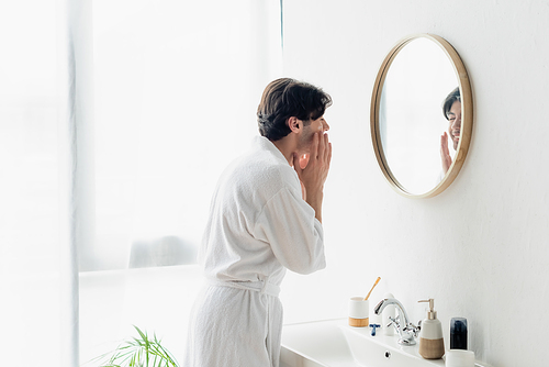 brunette man in white bathrobe applying face cream near mirror and sink