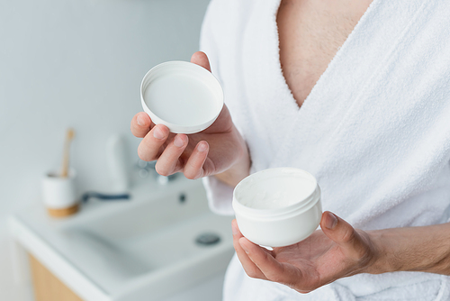 cropped view of man in white bathrobe holding cosmetic cream in blurred bathroom