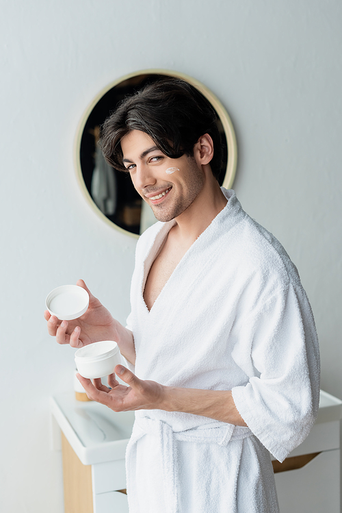 man in bathrobe smiling at camera while holding cosmetic cream in bathroom
