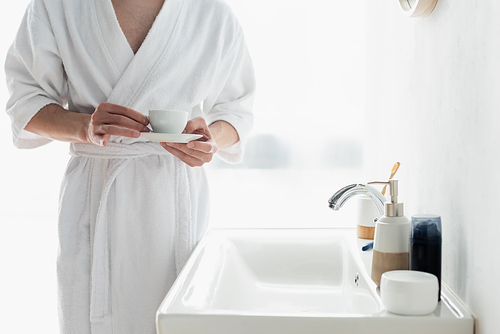 cropped view of man in white bathrobe holding coffee cup near sink and containers with toiletries