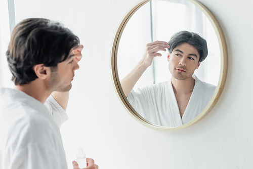 young man in white bathrobe applying cosmetic serum near mirror in bathroom