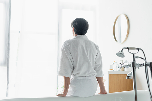back view of brunette man in white bathrobe sitting on bathtub
