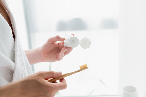 cropped view of man in white bathrobe holding toothbrush and tube of toothpaste