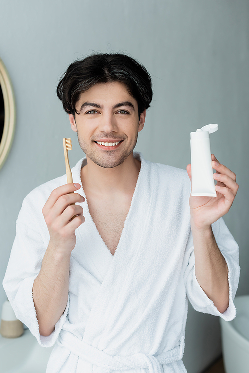 happy man with toothbrush and tube of toothpaste looking in camera in bathroom