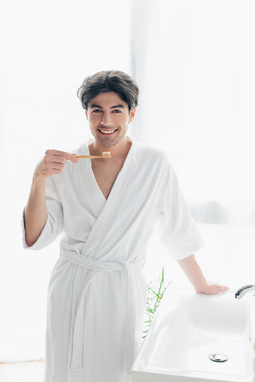 joyful man in white bathrobe  while holding toothbrush with toothpaste in bathroom
