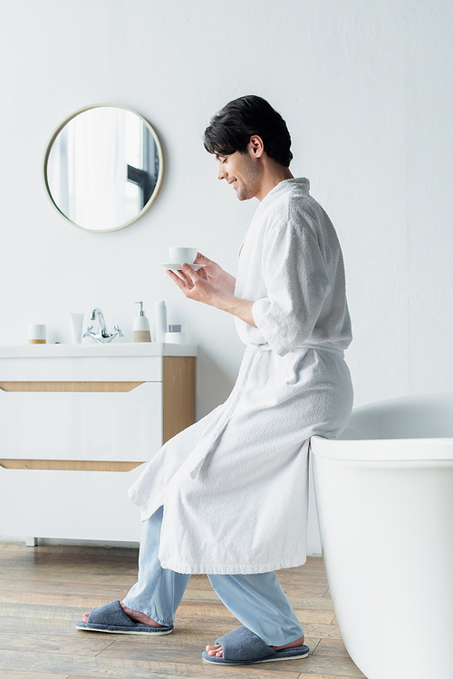side view of happy man in bathrobe sitting on bathtub with cup of coffee
