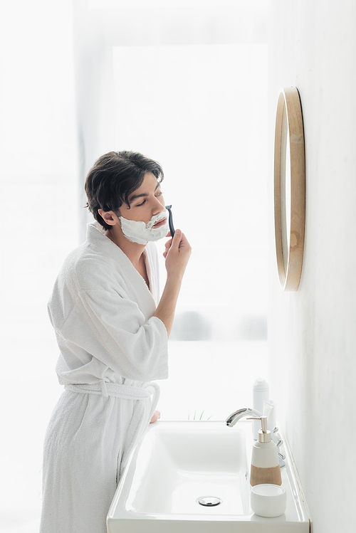 young man in white bathrobe shaving near mirror and sink in bathroom