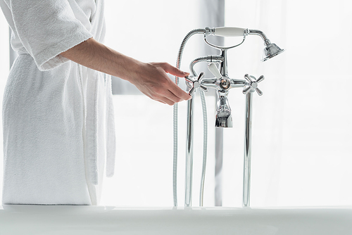 partial view of man in white bathrobe opening faucet near bathtub
