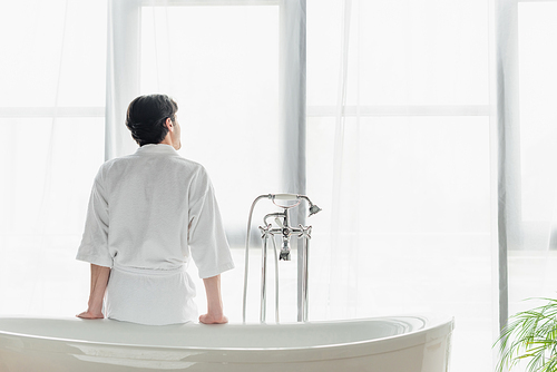 back view of man in white bathrobe sitting on bathtub near window