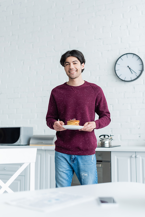 brunette man smiling at camera while holding tasty pancakes in kitchen