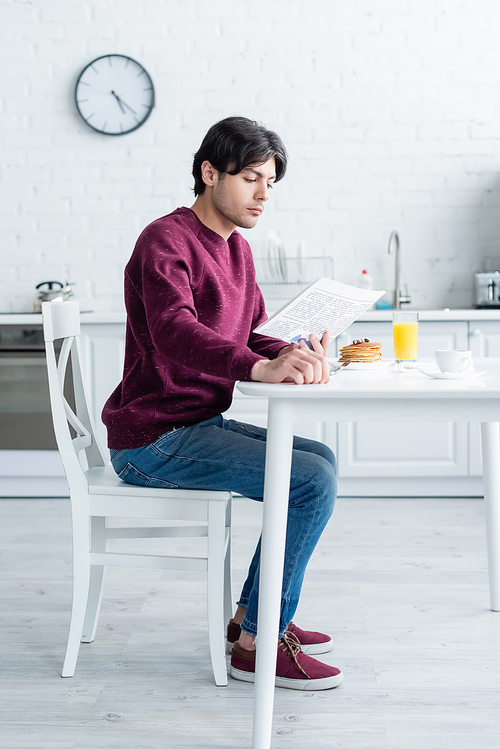 full length view of man reading morning newspaper near breakfast on kitchen table