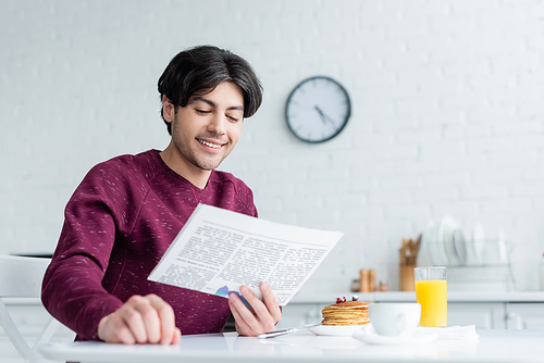 smiling man reading newspaper near pancakes and beverages on kitchen table