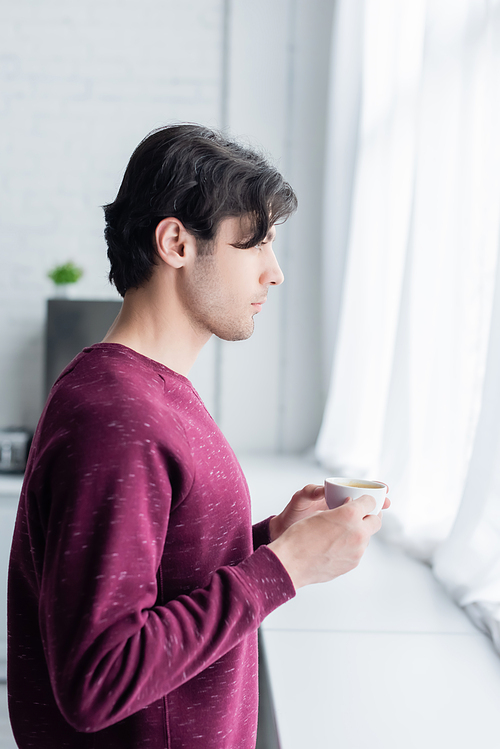 side view of young brunette man standing with coffee cup near window