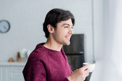 young man smiling while holding coffee cup in blurred kitchen