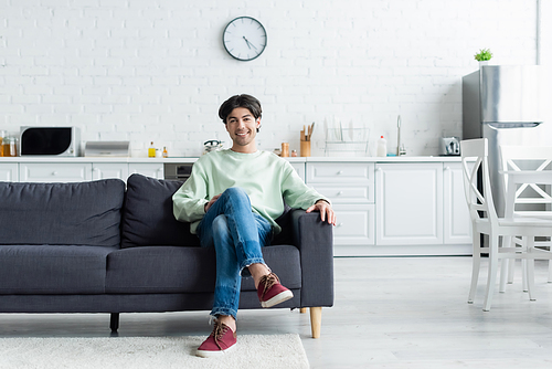 full length view of smiling man sitting on couch in modern kitchen with white furniture