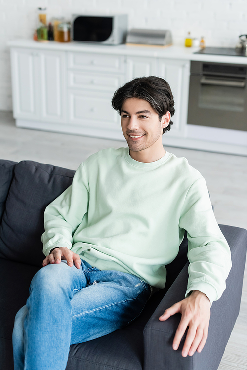 pleased brunette man sitting on sofa in blurred kitchen
