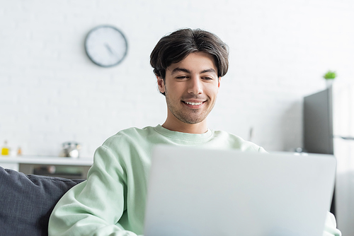 smiling brunette man working on blurred laptop at home