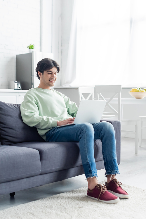 full length view of brunette man smiling while sitting on couch with laptop