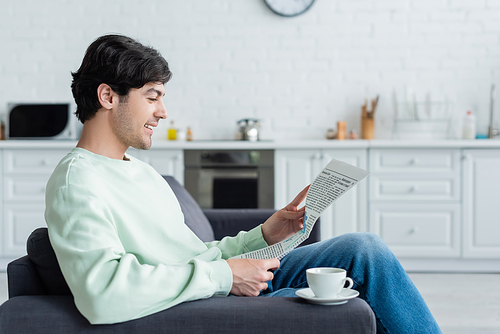 side view of smiling man reading morning newspaper on couch near coffee cup