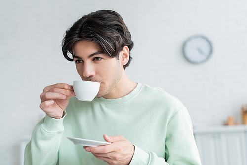 young brunette man holding saucer while drinking morning coffee