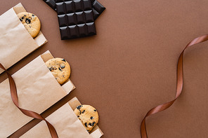 Top view of cookies in craft packages, chocolate bars and ribbon on brown background