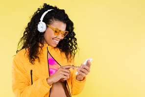 happy african american young woman in headphones using smartphone isolated on yellow