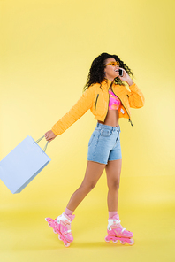 full length of african american young woman on pink roller skates talking on cellphone while holding shopping bag on yellow