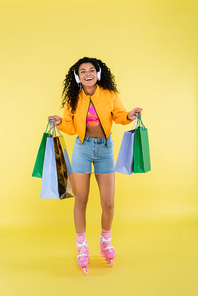 full length of happy african american young woman in roller skates and headphones holding shopping bags on yellow