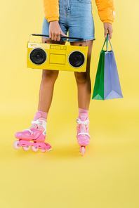 cropped view of african american woman in roller skates holding shopping bags and boombox on yellow