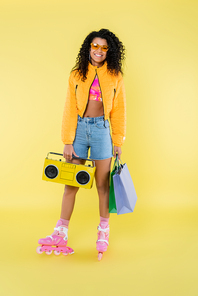 full length of happy african american woman in sunglasses and roller skates holding shopping bags and boombox on yellow