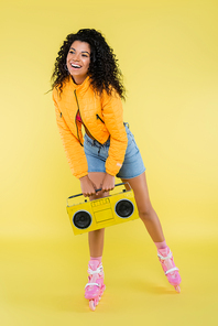 full length of cheerful african american woman on roller skates holding retro boombox on yellow