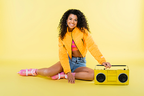 full length of smiling african american woman on roller skates sitting near retro boombox on yellow