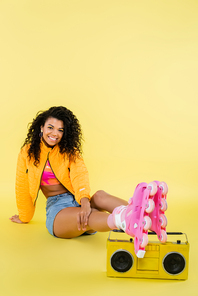 full length of cheerful african american woman on roller skates sitting near retro boombox on yellow