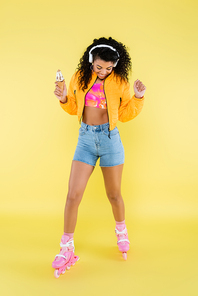 happy african american young woman in headphones and roller skates holding ice cream cone on yellow