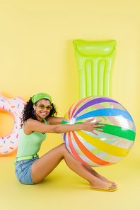 full length of joyful african american woman sitting with inflatable ball, mattress and ring on yellow