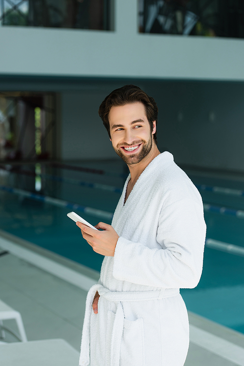 Positive man in bathrobe holding smartphone in spa center