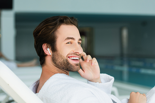 Smiling man in wireless earphone and bathrobe relaxing on deck chair in spa center