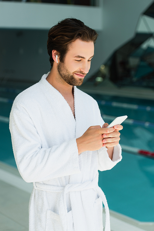Young man in wireless earphone and bathrobe using smartphone in spa center