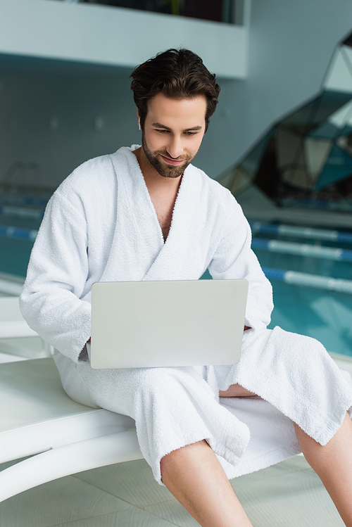 Young man in bathrobe and earphone using laptop in spa center