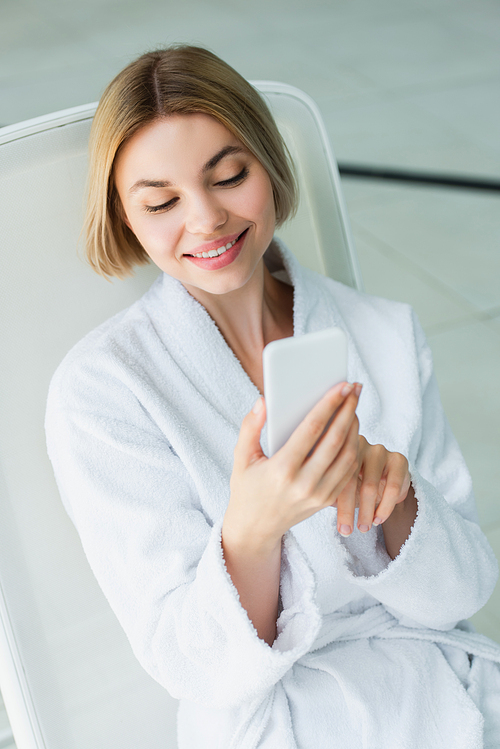 Smiling woman in bathrobe using smartphone on deck chair in spa center