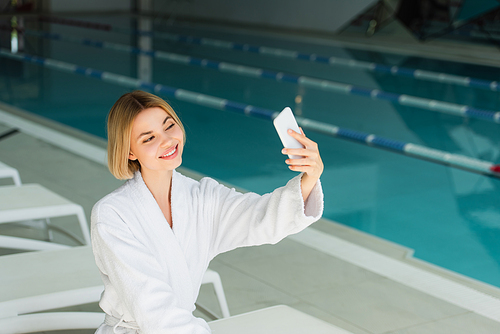 Smiling woman taking selfie on smartphone near swimming pool in spa center