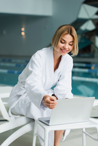 Cheerful woman in bathrobe using laptop on deck chair in spa center