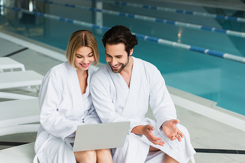 Smiling couple in bathrobes using laptop on deck chair in spa center