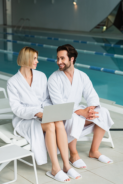 Young couple in bathrobes smiling at each other while using laptop in spa center