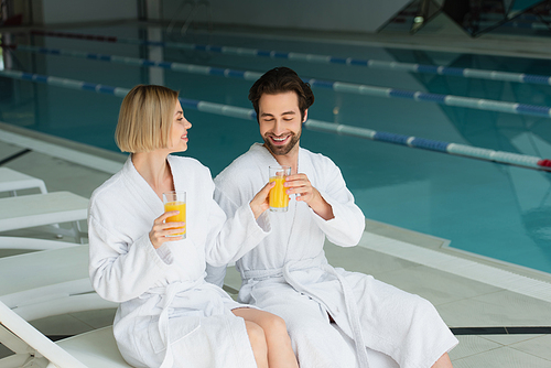 Smiling woman in bathrobe giving orange juice to boyfriend in spa center