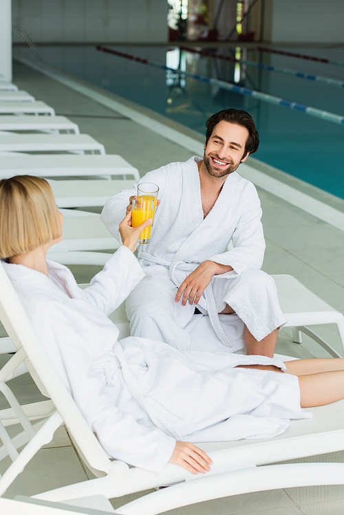 Smiling man clinking orange juice with girlfriend on deck chair in spa center