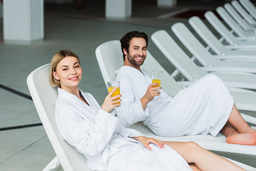 Young woman holding orange juice near blurred boyfriend on deck chair in spa center