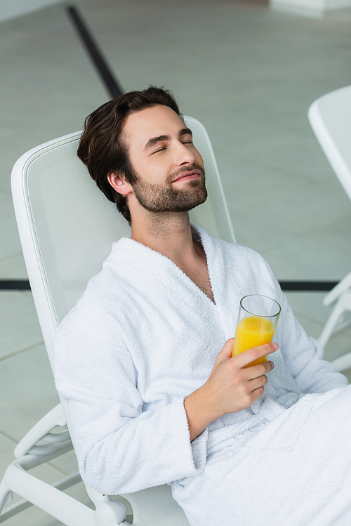 Young man in white bathrobe holding orange juice while relaxing in spa center
